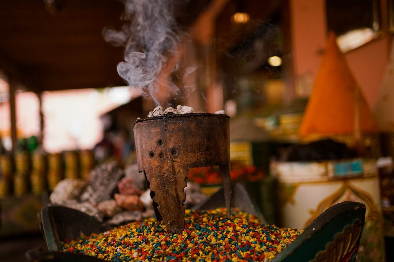 Close-up of a rusted incense burner emitting smoke over colorful spices in a market setting.